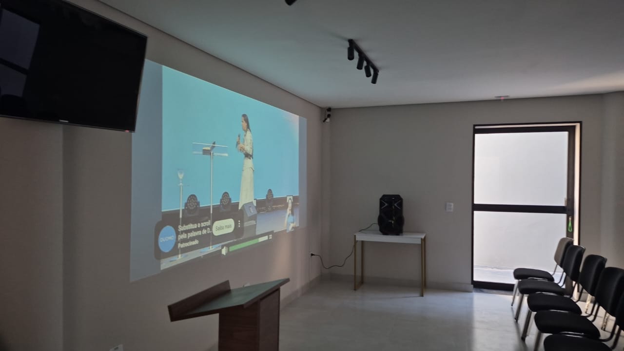 Parede com projeção em tela grande, púlpito de madeira, caixa de som e televisor.